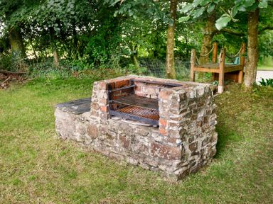 An outdoor stone barbecue grill on grass with trees and a wooden bench in the background at The Roundhouse Warbstow