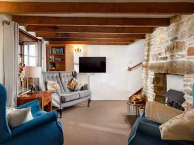 A living room with wooden beams a stone fireplace two blue armchairs a grey sofa a wooden side table a lamp bookshelves and a wall-mounted TV at Ash Cottage in Warbstow