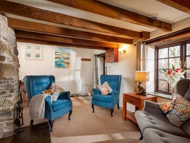 A sitting room with blue armchairs wooden beams stone wall and a window with flowers at Ash Cottage in Warbstow