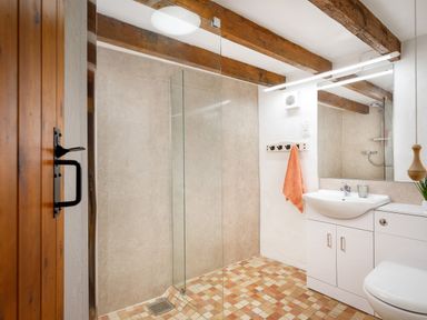 A bathroom with a walk-in shower with glass screen a white sink with cabinet a large mirror wooden ceiling beams and a tiled floor at Ash Cottage in Warbstow