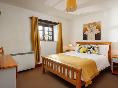 A bedroom with a wooden bed a desk and a window with yellow curtains at Ash Cottage in Warbstow