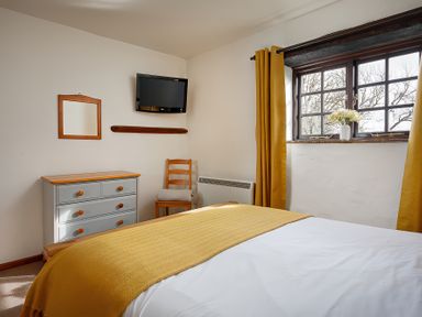 A bedroom with a bed covered by a yellow blanket and white sheets a dresser a wooden chair with a folded blanket a wall-mounted TV and a window with yellow curtains and flowers on the sill at Ash Cottage in Warbstow