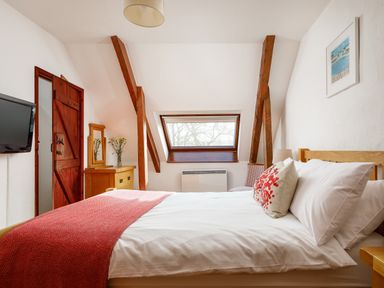 A bedroom with a bed with red and white bedding a wooden dresser with a mirror a wall-mounted television a window and exposed wooden beams at Pond Meadow in Warbstow