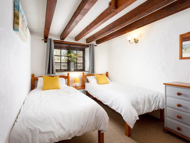 A bedroom with two single beds white bedding yellow pillows a window with curtains and flower vase and wooden beams on ceiling at Pond Meadow in Warbstow