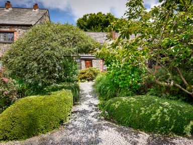 A garden path with neatly trimmed bushes and a large arching shrub leading to a stone house at Pond Meadow in Warbstow