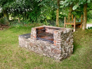 An outdoor stone barbecue grill with metal grates and a wooden bench among trees at Pond Meadow in Warbstow