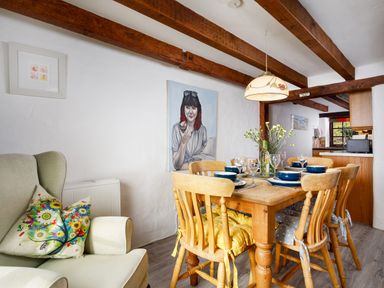 A dining area with wooden table and chairs with cushions next to an armchair with decorative pillows and artwork on walls at Little Leighs Warbstow