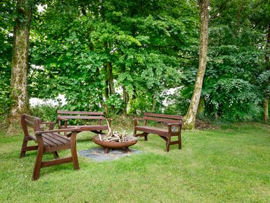 A circle of wooden benches around a metal fire pit with logs on a grass lawn by trees at Little Leighs in Warbstow