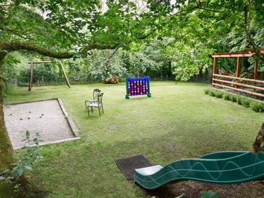 A grassy backyard with a blue connect four game two black metal chairs a wooden swing set a bocce ball court and a green slide at Little Leighs in Warbstow