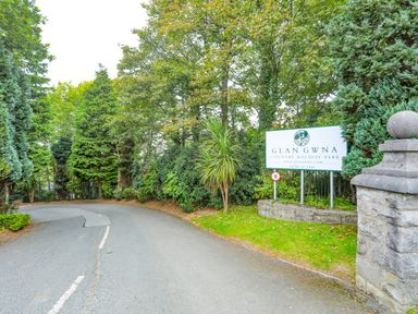 A road curving left with a sign for Glan Gwna Country Holiday Park surrounded by trees and greenery at Eirlys in Caeathro near Caernarfon