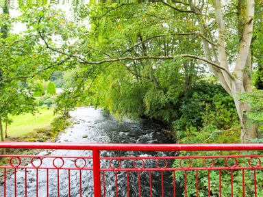 A river flowing under a red railing with trees on the riverbanks at Eirlys in Caeathro near Caernarfon