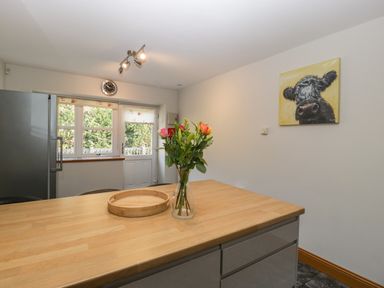 A kitchen with a wooden counter with a vase of flowers a round wooden tray a refrigerator a clock above a window and a door and a painting of a cow on the wall at About Time Cottage in Parkend
