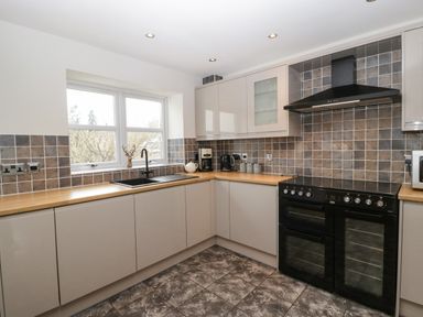 A kitchen with tiled backsplash, black stove, sink under a window, and beige cabinets at About Time Cottage in Parkend