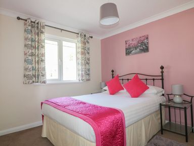 A bedroom with a double bed with a white duvet and pink cushions a pink wall with a tree picture a window with floral curtains and bedside tables with lamps