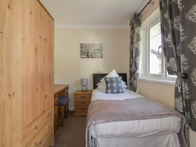 A bedroom with a single bed a wooden wardrobe bedside table with lamp and a window with patterned curtains at Watergate Bay in Liskeard
