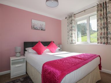 A bedroom with a double bed with pink pillows and bed runner beside a window with floral curtains at Watergate Bay in Liskeard
