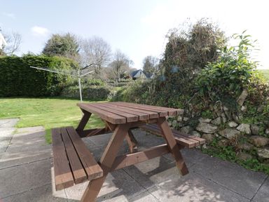 A wooden picnic table on a paved patio next to a grassy yard with bushes and trees at Watergate Bay in Liskeard