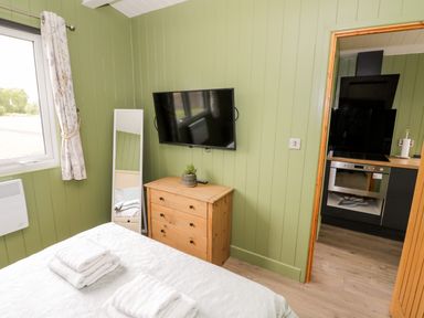 A bedroom with green walls a wooden chest of drawers a wall-mounted TV and a doorway to a kitchen at Cotswold View 1 in Honeybourne