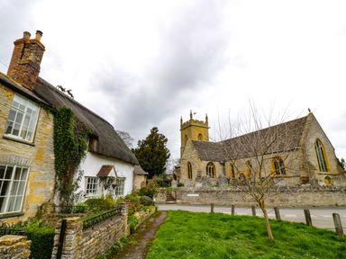 A stone cottage with a thatched roof and a church with a tower in the background in a village setting at Cotswold View 1 in Honeybourne