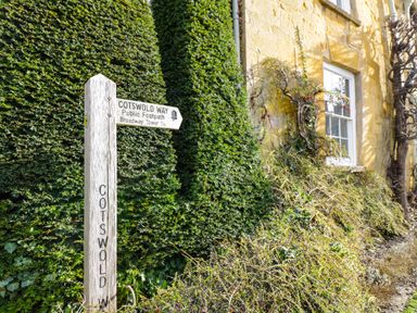 A wooden Cotswold Way signpost next to trimmed bushes and a yellow stone building with a window at Cotswold View 1 in Honeybourne