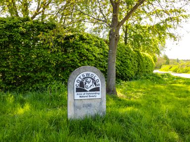 A stone sign reading COTSWOLDS Area of Outstanding Natural Beauty beside a tree and hedge in a grassy area at Cotswold View 1 in Honeybourne