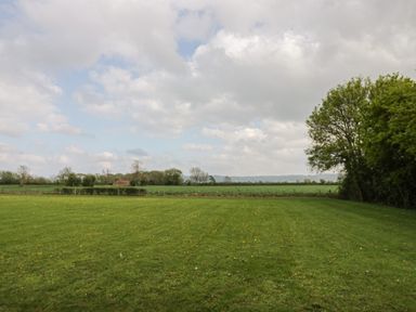 A grassy field with scattered yellow flowers trees on the right and distant farmland under a cloudy sky at Cotswold View 1 in Honeybourne