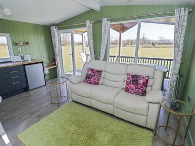 A living room with a white sofa with floral pillows green walls a small kitchenette and large windows at Cotswold View 1 in Honeybourne