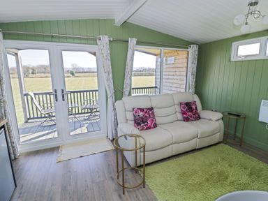 A living room with a cream sofa and glass tables next to windows overlooking a balcony and fields at Cotswold View 1 in Honeybourne