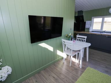 A small dining area with a white table and two chairs a large mounted TV and a kitchen with a window in Cotswold View 1 in Honeybourne