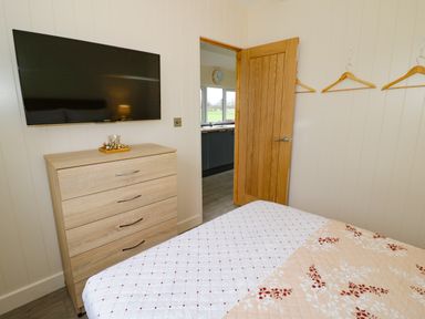 A bedroom with a wooden dresser and wall mounted tv near an open door and three wooden hangers on the wall at Cotswold View 2 in Honeybourne