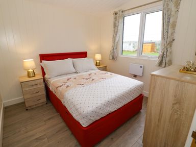 A bedroom with a red bed frame bedside tables with lamps a wooden dresser and a window with curtains at Cotswold View 2 in Honeybourne