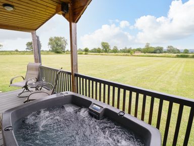 An outdoor covered wooden deck with a hot tub two chairs and a small table overlooking a large grassy field at Cotswold View 2 in Honeybourne