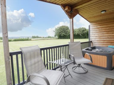 A covered wooden deck with two chairs a small glass table with glasses and a hot tub overlooking a grass field at Cotswold View 2 in Honeybourne