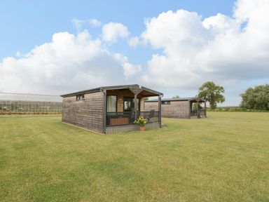 Two wooden cabins on a large grassy field under a cloudy sky at Cotswold View 2 in Honeybourne
