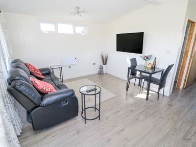 A living room with a black sofa with red pillows a glass coffee table a small dining table with two chairs a wall-mounted tv and light wood flooring at Cotswold View 2 in Honeybourne
