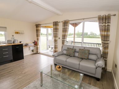 A living room with a gray sofa and glass coffee table next to a kitchen area with a small fridge at Cotswold View 3 in Bretforton near Evesham
