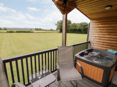 A covered wooden deck with two chairs a small glass table with two wine glasses and a hot tub overlooking a grassy field at Cotswold View 3 Bretforton near Evesham
