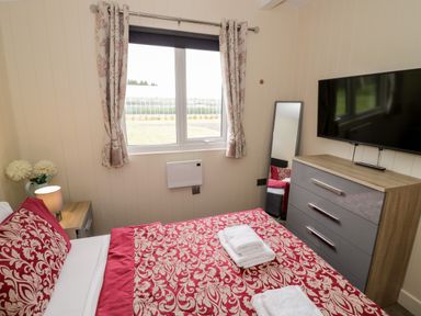 A bedroom with a bed covered in red and beige patterned bedding a chest of drawers with a TV on top and a standing mirror near a window with curtains at Cotswold View 4 Bretforton near Evesham
