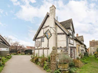 A rustic inn building with a sign hanging on the wall and garden plants along a stone fence at Cotswold View 4 in Bretforton near Evesham