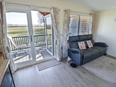 A living area with a dark sofa and patterned cushions near glass doors opening to a balcony with chairs at Cotswold View 4 Bretforton near Evesham