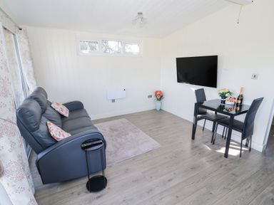 A living room with a sofa, small table, wall-mounted TV, dining table with two chairs, and flowers at Cotswold View 4 Bretforton near Evesham