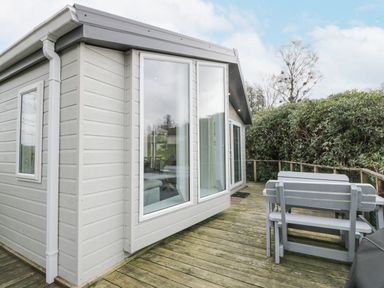 An outdoor wooden deck with a gray picnic table and a small house with large windows at Oyster Bay Lodge in Dalbeattie