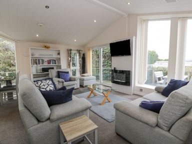 A living room with grey sofas and blue cushions a glass coffee table a wall mounted television and large windows at Oyster Bay Lodge in Dalbeattie