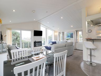 A dining area with white chairs and glass table next to a living room with grey sofas and a wall-mounted TV at Oyster Bay Lodge in Dalbeattie