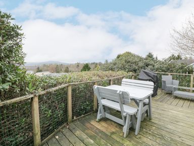 An outdoor wooden deck with a table and benches a barbecue grill and chairs surrounded by greenery at Oyster Bay Lodge in Dalbeattie