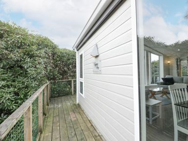 A wooden deck with railing outside a light gray building with windows showing a living room at Oyster Bay Lodge in Dalbeattie