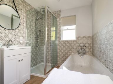 A bathroom with a white sink cabinet a round mirror a glass shower enclosure and a white bathtub with patterned tiles on the walls at Tresawle in Crantock