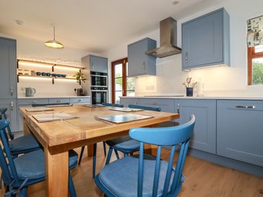 A kitchen with blue cabinets, wooden dining table, blue chairs, shelves with glasses and cups, and a door at Tresawle in Crantock