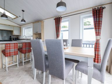 A dining area with a wooden table and grey chairs next to windows with red plaid curtains and a kitchen counter with red plaid bar stools at Beachcliff in Porth
