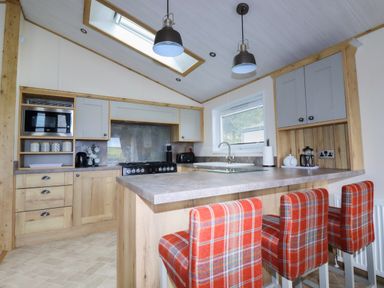 A kitchen with wooden cabinets a countertop with three red plaid chairs a window and two hanging lamps at Beachcliff in Porth
